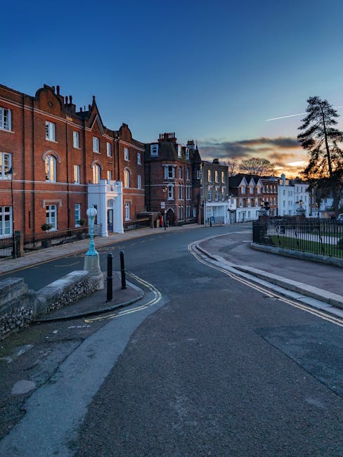 A quiet residential street with multi-story brick and stucco buildings, featuring small windows and decorative architectural elements. The scene shows a paved road with curbs, a lamppost, and black bo