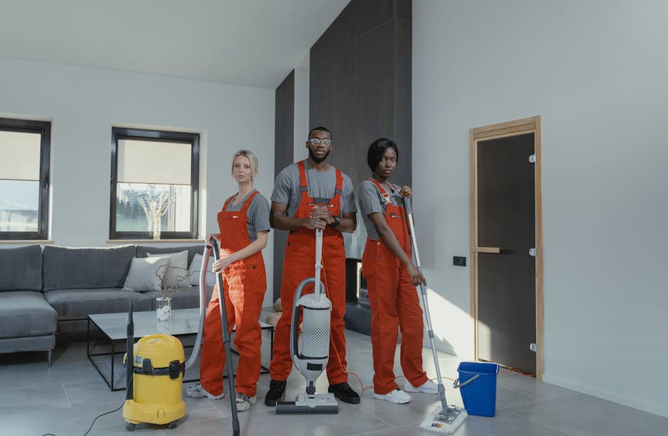 Two professional cleaners from Harrow Cleaner are performing surface cleaning and deep cleaning in a modern living room. One cleaner is using a vacuum cleaner to clean the wooden floor near a sofa, wh