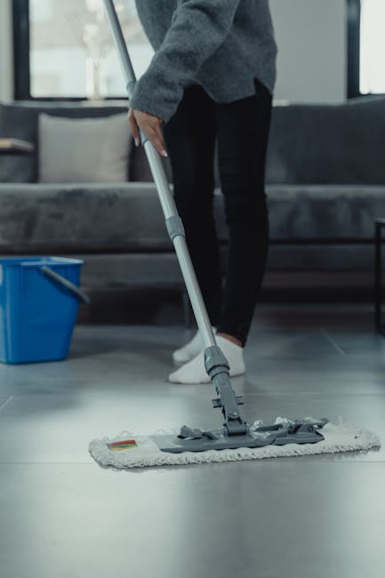 A person wearing dark clothing and footwear is using a mop to clean a smooth, polished floor in a modern indoor setting. The mop has a flat, rectangular cleaning head with a white cloth attached, and