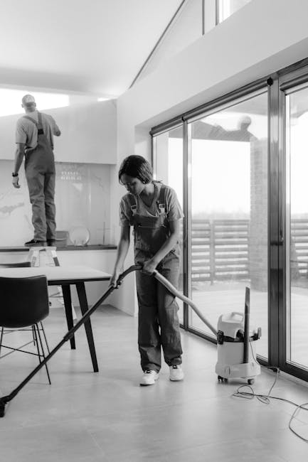 A professional cleaner from Harrow Cleaner is performing surface cleaning in a modern, well-lit dining area. The woman is vacuuming a light-colored wooden floor with a vacuum cleaner, while a man on a step stool in the background appears to be working on the wall or ceiling. The room features large sliding glass doors allowing natural light to fill the space, with a sitting area visible beyond. The surfaces, including the polished wooden floor and glass doors, appear clean and dust-free, reflecting a high standard of deep cleaning and sanitisation. The environment looks neat and well-maintained, showcasing the company's expertise in domestic cleaning services.
