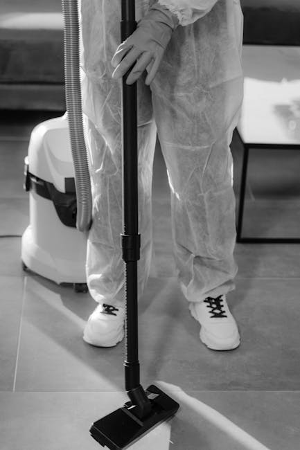 A professional cleaner from Harrow Cleaner is performing surface cleaning in a modern, well-lit dining area. The woman is vacuuming a light-colored wooden floor with a vacuum cleaner, while a man on a step stool in the background appears to be working on the wall or ceiling. The room features large sliding glass doors allowing natural light to fill the space, with a sitting area visible beyond. The surfaces, including the polished wooden floor and glass doors, appear clean and dust-free, reflecting a high standard of deep cleaning and sanitisation. The environment looks neat and well-maintained, showcasing the company's expertise in domestic cleaning services.