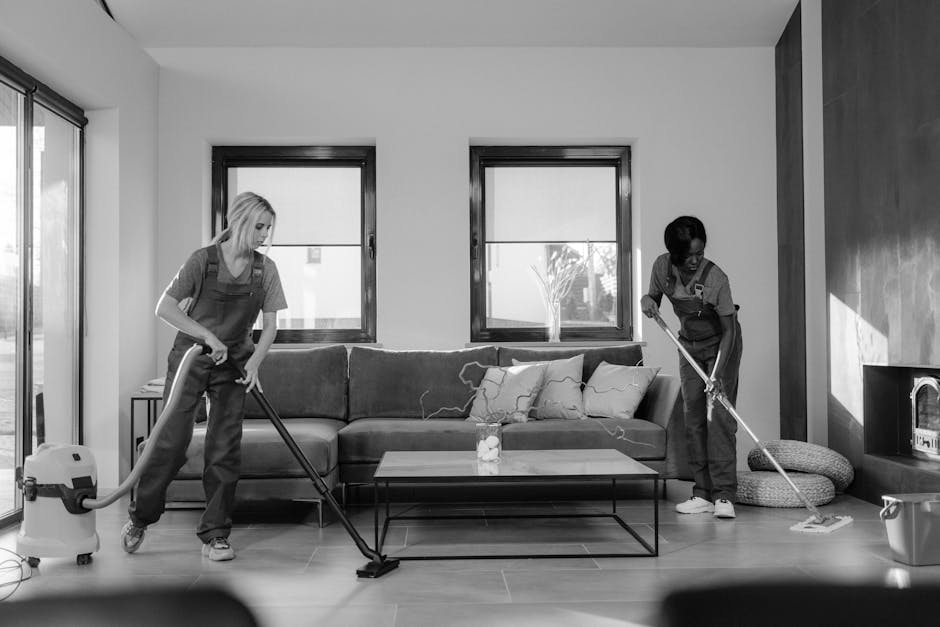 Two professional cleaners from Harrow Cleaner are engaged in surface cleaning within a modern kitchen. One cleaner, a woman, is using a vacuum cleaner and appears to be thoroughly cleaning the tiled f