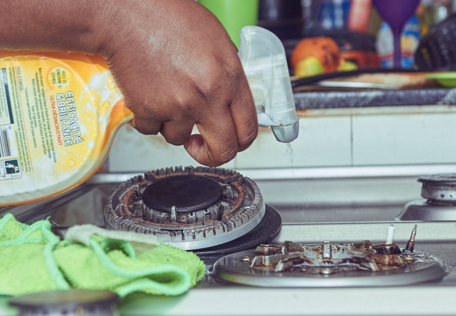 A woman with blonde hair tied back, wearing a white t-shirt, black apron, yellow and blue rubber cleaning gloves, is performing surface cleaning in a kitchen. She is using a cloth and spray bottle to 